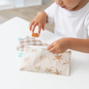A child in a white shirt holds Bumkins Reusable Snack Bag, Small 2 Pack: Palm Check while reaching for a snack at a white table.