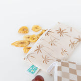 Banana chips spill from a Bumkins Reusable Snack Bag, Small 2 Pack: Palm Check on a white background.