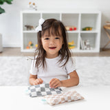 A smiling girl with a white bow holds a Bumkins Reusable Snack Bag, Small 2 Pack: Double Check, at a white table with a shelf behind her.