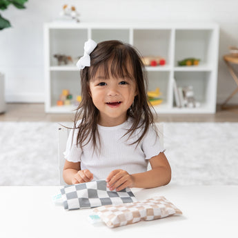 A smiling girl with a white bow holds a Bumkins Reusable Snack Bag, Small 2 Pack: Double Check, at a white table with a shelf behind her.
