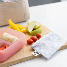 Load image into Gallery viewer, Cherry tomatoes spill from a Roamio by Bumkins Reusable Snack Bag, Small 2 Pack: Disco Boots on a cutting board near a bento box and fruit.
