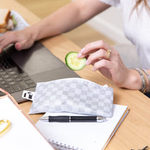 Load image into Gallery viewer, A person on a laptop holds a cucumber slice over a Roamio by Bumkins Reusable Snack Bag, Small 2 Pack: Rich &amp; Boujee by a notepad &amp; pen on a desk.
