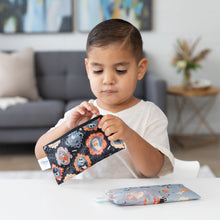 Load image into Gallery viewer, A young child clutches a Bumkins Reusable Snack Bag, Small 2 Pack: Everyday Is Halloween at a table, with a couch and plant in the background.
