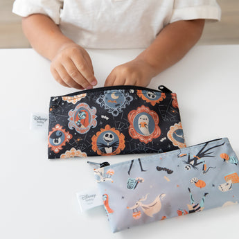 A child reaches into a Bumkins Reusable Snack Bag, Small 2 Pack: Everyday Is Halloween on a white table with another bag nearby.