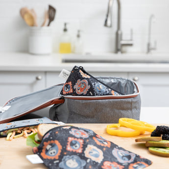 A Bumkins Reusable Snack Bag, Small 2 Pack: Everyday Is Halloween sits in a gray lunch bag on a kitchen counter, surrounded by food.