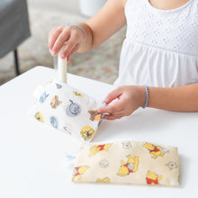 Load image into Gallery viewer, A child in a white top places an item into a Bumkins Reusable Snack Bag, Small 2 Pack: Pooh Bear and Friends on a white table.
