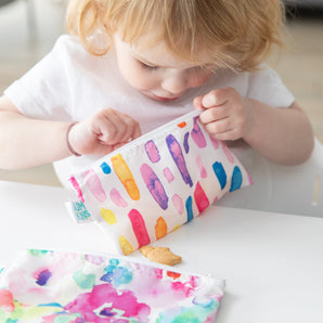A child looks in a Bumkins Reusable Snack Bag, Small 2-Pack: Watercolor & Brush Strokes while another bag sits on a white table.