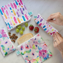 Load image into Gallery viewer, Hands zip a Bumkins Reusable Snack Bag, Small 2-Pack: Watercolor &amp; Brush Strokes near other matching bags and snacks on a marble surface.
