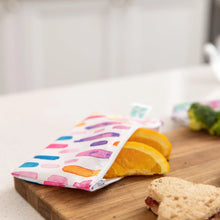 Load image into Gallery viewer, A Bumkins Reusable Snack Bag, Small 2-Pack: Watercolor &amp; Brush Strokes holds orange slices on a cutting board next to a sandwich and broccoli.
