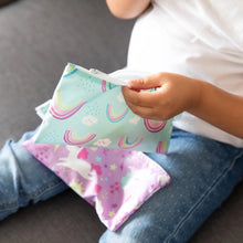 Load image into Gallery viewer, Close-up of a child on a gray couch holding a Bumkins Reusable Snack Bag, Small 2-Pack: Rainbows &amp; Unicorns with another on their lap.
