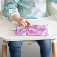 Load image into Gallery viewer, Close-up of a child in a rainbow bib with a purple Bumkins Reusable Snack Bag, Small 2-Pack: Rainbows &amp; Unicorns on a high chair tray.
