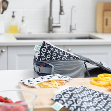Load image into Gallery viewer, A Bumkins Reusable Snack Bag, Small 2-Pack: XOXO &amp; Hearts sits in an open lunch box while the other bag sits nearby on the kitchen counter.
