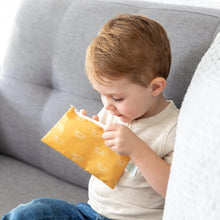 Load image into Gallery viewer, A child on a gray couch peers into a yellow Bumkins Reusable Snack Bag, Small 2-Pack: Sunshine and Grounded.
