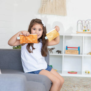 A girl smiles on a gray couch, holding two bags from Bumkins Reusable Snack Bag 4 Pack: Desert Boho.