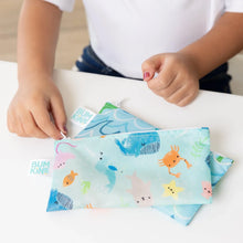 Load image into Gallery viewer, Close-up of a child zipping a Bumkins Reusable Snack Bag, Small 2-Pack: Ocean Life &amp; Whale Tail on top of another bag on a white table.
