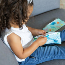Load image into Gallery viewer, A curly-haired child, in a white top and blue jeans, holds a Bumkins Reusable Snack Bag, Small 2-Pack: Ocean Life &amp; Whale Tail on a couch.
