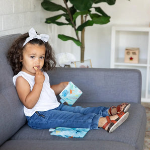 A toddler with curly hair and a bow enjoys snacks from Bumkins Reusable Snack Bag, Small 2-Pack: Ocean Life & Whale Tail on a gray sofa.