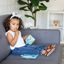 Load image into Gallery viewer, A toddler with curly hair and a bow enjoys snacks from Bumkins Reusable Snack Bag, Small 2-Pack: Ocean Life &amp; Whale Tail on a gray sofa.
