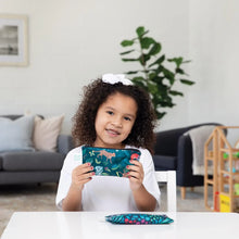 Load image into Gallery viewer, A curly-haired child holds a Bumkins Reusable Snack Bag, Small 2-Pack: Jungle &amp; Animal Prints with another bag resting on a white table.
