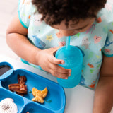 A toddler in a bib drinks from a Bumkins Silicone Straw Cup with Lid: Blue at a white table beside a blue plate of food.