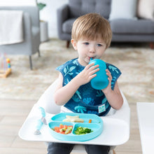 Load image into Gallery viewer, A child in a high chair wearing a blue bib drinks from a Bumkins Silicone Straw Cup with Lid: Blue, with a blue plate of food nearby.
