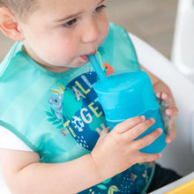 Load image into Gallery viewer, A toddler uses a Bumkins Silicone Straw Cup with Lid: Blue, drinking independently indoors while wearing a bib.
