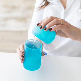 A person at a white table pours water into a Bumkins Silicone Straw Cup with Lid: Blue using another cup.