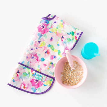 A folded colorful bib, a pink bowl of cereal, a matching spoon, and a Bumkins Silicone Straw Cup with Lid: Blue on a white background.