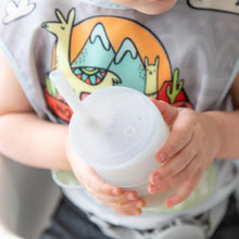 Load image into Gallery viewer, A child proudly holds a Bumkins Silicone Straw Cup with Lid: Gray while wearing a dinosaur bib.
