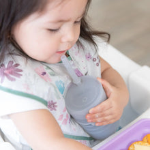 Load image into Gallery viewer, A toddler with a floral bib sits at a table, holding a Bumkins Silicone Straw Cup with Lid: Gray, exploring independent drinking.
