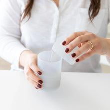 Load image into Gallery viewer, In a white shirt, a person uses a cup to pour water into a Bumkins Silicone Straw Cup with Lid: Gray at a white table.

