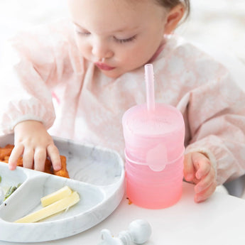 A toddler in a light pink bib eats from a gray sectioned plate with a Bumkins Silicone Straw Cup with Lid: Pink nearby at a white table.