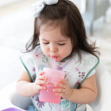 Load image into Gallery viewer, A toddler in a floral bib sips from a Bumkins Silicone Straw Cup with Lid: Pink, with a bow in her hair.
