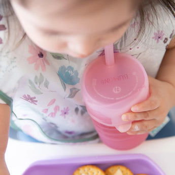 A toddler confidently drinks from a Bumkins Silicone Straw Cup with Lid: Pink, while gazing at a purple tray of crackers. 