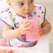 Load image into Gallery viewer, A baby with a colorful bib sips from a Bumkins Silicone Straw Cup with Lid: Pink in a high chair.
