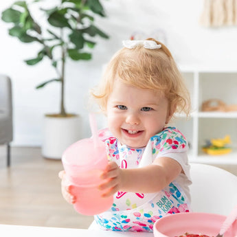 A smiling toddler in a high chair holds a Bumkins Silicone Straw Cup with Lid: Pink, with a plant and shelves in the background.
