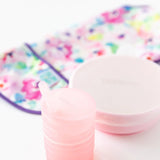 Bumkins Silicone Straw Cup with Lid: Pink sits on a white background, with a pink bowl and floral bib blurred behind.