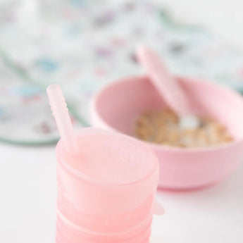 Close-up of Bumkins Silicone Straw Cup with Lid: Pink on a white background, with a pink bowl of cereal and gray bib blurred behind.