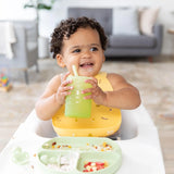 A baby in a high chair, wearing a yellow bib, holds a Bumkins Silicone Straw Cup with Lid: Sage, with food on a divided plate in front.