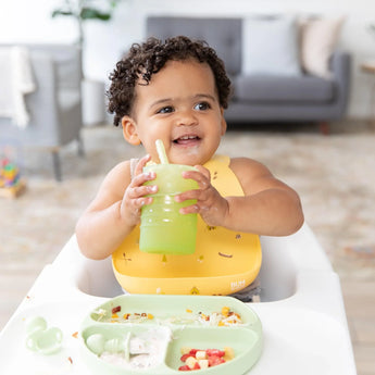 A baby in a high chair, wearing a yellow bib, holds a Bumkins Silicone Straw Cup with Lid: Sage, with food on a divided plate in front.