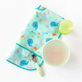 Bumkins Silicone Straw Cup with Lid: Sage sits near a folded ocean-themed bib, a green bowl with lid, & a green spoon on a white background.