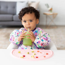 Load image into Gallery viewer, A toddler with a colorful bib sips from a Bumkins Silicone Straw Cup with Lid: Sage beside a pink mat with chopped fruits and veggies.
