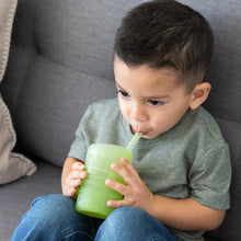 Load image into Gallery viewer, A child in a gray shirt sits on a couch, sipping from a Bumkins Silicone Straw Cup with Lid: Sage.
