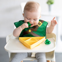 Load image into Gallery viewer, A baby in a Bumkins Caped SuperBib®: The Legend of Zelda™ Link sits in a high chair, eating from a yellow divided plate with green utensils.
