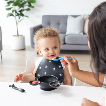 A smiling toddler in a bib is fed by an adult with a Bumkins Silicone Dipping Spoons: Mickey Mouse (Black and Blue), while a bowl sits nearby.