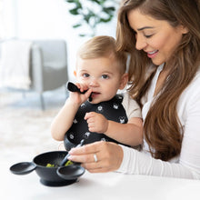 Load image into Gallery viewer, A woman smiles as a baby chews a Bumkins Silicone Dipping Spoons: Mickey Mouse (Classic Black), with a black bowl of food on a table.
