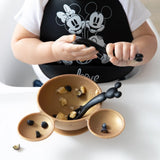 A child in a black bib holds a Bumkins Silicone Dipping Spoons: Mickey Mouse (Classic Black), with a gold bowl of food on a high chair tray.