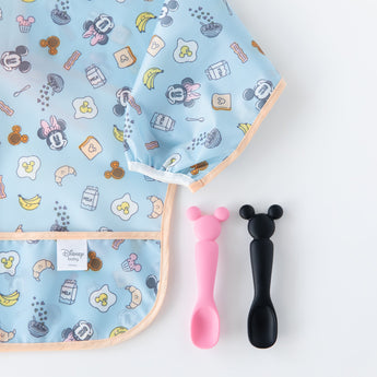 Bumkins Silicone Dipping Spoons: Minnie Mouse (Black and Pink) sit next to a blue sleeved bib with breakfast pattern on a white background.