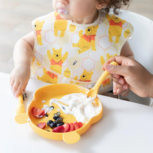 Load image into Gallery viewer, A child enjoys yogurt with fruit on a bear-shaped plate, using Bumkins Silicone Dipping Spoons: Winnie the Pooh with adult assistance.
