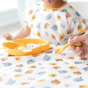 A child enjoys yogurt with Bumkins Silicone Dipping Spoons: Winnie the Pooh from a yellow plate on a tablecloth in a patterned bib.
